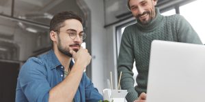 Portrait of friendly comapny workers look attentively at laptop computer, sit at office desk, happy to succeed in creating business project, eats healthy and unhealthy food. People, dinner time, work